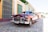 A classic vintage car with a shiny, burgundy exterior is parked on a cobblestone street, reflecting the surrounding colorful buildings. The car has chrome detailing and a visible license plate. There are pedestrians walking by, and the scene is set under a bright, clear sky.