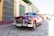 A classic vintage car with a shiny, burgundy exterior is parked on a cobblestone street, reflecting the surrounding colorful buildings. The car has chrome detailing and a visible license plate. There are pedestrians walking by, and the scene is set under a bright, clear sky.