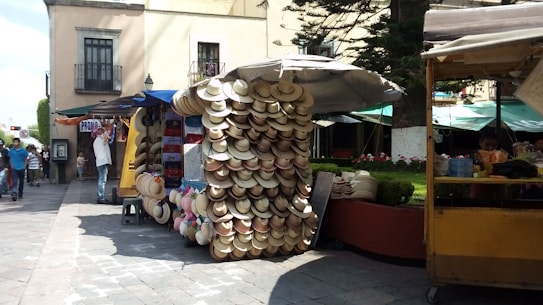 A street market stall displaying a large assortment of hats hanging and arranged neatly. The surrounding area shows people walking, an ATM, and nearby booths with items and refreshments. A quaint backdrop with buildings and trees adds to the market setting.