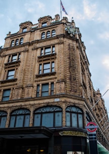 A large, ornate building with detailed stone architecture adorned with rows of windows and decorative elements. A British flag flies from the top, and below, there is a sign for the London Underground with a station name visible. The sky is slightly overcast.