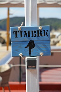 A wooden sign with the word 'TIMBRE' in black stencil lettering is mounted on a white post. The sign is painted in a light blue color and features a black hand pointing downwards. It is attached to the post with knotted ropes. Below the sign, there is a modern doorbell device attached to the post.