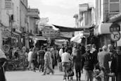 A crowded street market features numerous individuals engaged in browsing and shopping. Various signs and storefronts line the narrow street, with awnings providing some shade. Bicycles are visible on the left side, while groups of people walk in both directions. A stop sign in Turkish is visible on the right.