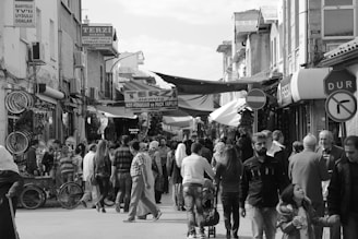 A crowded street market features numerous individuals engaged in browsing and shopping. Various signs and storefronts line the narrow street, with awnings providing some shade. Bicycles are visible on the left side, while groups of people walk in both directions. A stop sign in Turkish is visible on the right.