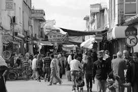 A crowded street market features numerous individuals engaged in browsing and shopping. Various signs and storefronts line the narrow street, with awnings providing some shade. Bicycles are visible on the left side, while groups of people walk in both directions. A stop sign in Turkish is visible on the right.