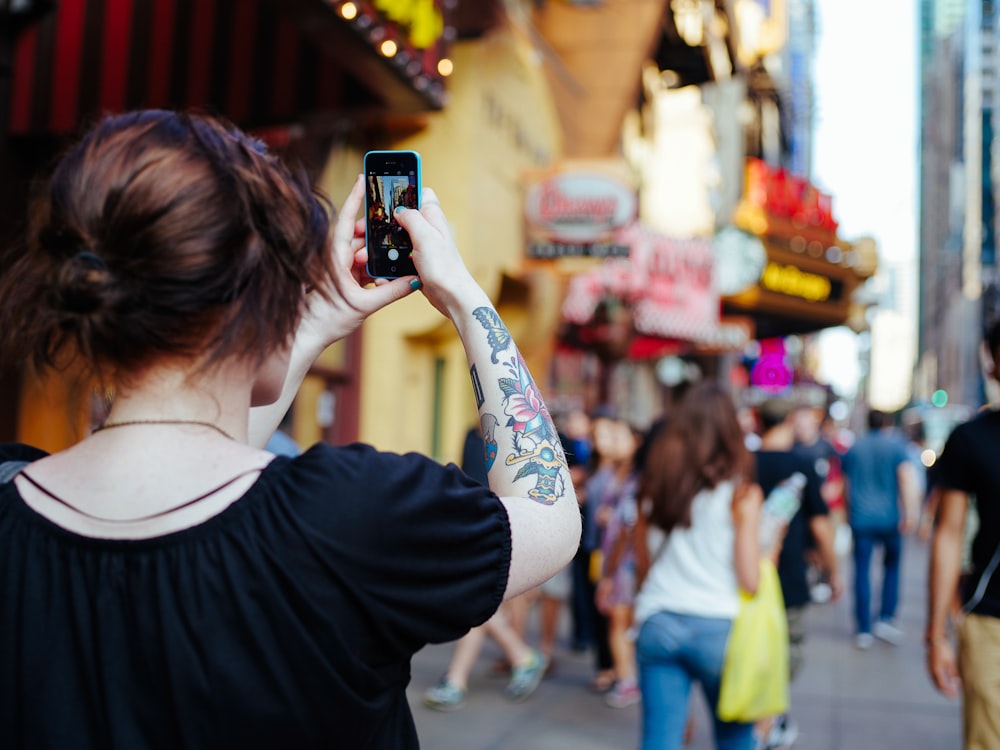 Fokusfoto einer Frau in schwarzem Hemd mit Kappenärmeln, die beim Fotografieren ein Smartphone in der Hand hält