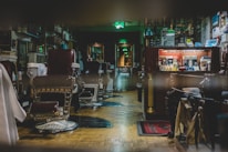 A vintage barbershop interior featuring antique barber chairs with red leather upholstery and metal frames. The shop is warmly lit and filled with various barber tools, products, and framed pictures on the walls. A counter displays hair products, brushes, and shaving supplies. Wooden flooring adds to the classic ambiance of the space.