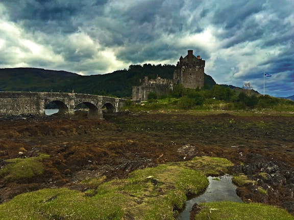 A historic stone castle stands proudly on a lush green landscape, surrounded by green shrubbery. A stone arched bridge extends over a body of water leading towards the castle. The background features rolling hills under a dramatic, overcast sky. A flag with a blue and white design flutters in the breeze.