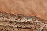 A partially eroded brick wall with exposed bricks beneath a layer of worn plaster. The wall shows a gradient of colors ranging from deep browns and reds to lighter beige and tan. The texture is rough with clear distinctions between the bricks and the plaster.