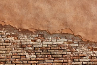 A partially eroded brick wall with exposed bricks beneath a layer of worn plaster. The wall shows a gradient of colors ranging from deep browns and reds to lighter beige and tan. The texture is rough with clear distinctions between the bricks and the plaster.