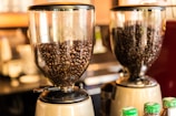 Two coffee grinders filled with dark roasted coffee beans, standing on a counter. The background is blurred, creating a warm and inviting atmosphere typical of a coffee shop.