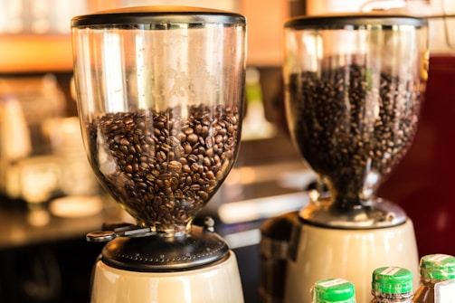 Two coffee grinders filled with dark roasted coffee beans, standing on a counter. The background is blurred, creating a warm and inviting atmosphere typical of a coffee shop.