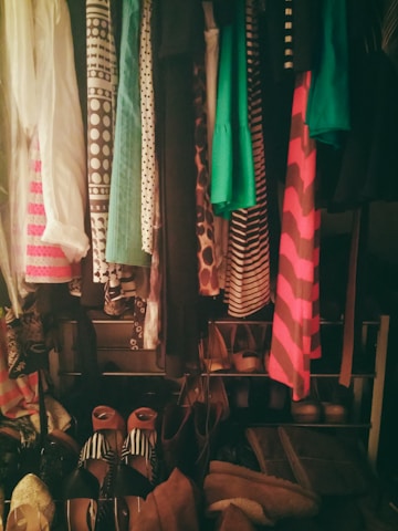 A closet filled with a variety of clothing items and shoes. The clothes are organized on hangers, displaying different patterns such as polka dots and stripes in colors like green, pink, and black. The bottom part shows an assortment of shoes arranged in pairs, with various designs including striped, open-toed, and heeled footwear.