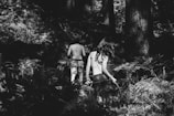 A black and white photograph of two children walking away into a dense forest. Both are bare-backed with patterns painted on their backs, surrounded by tall trees and ferns. The light and shadow interplay creates a mysterious and adventurous atmosphere.