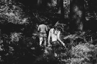 A black and white photograph of two children walking away into a dense forest. Both are bare-backed with patterns painted on their backs, surrounded by tall trees and ferns. The light and shadow interplay creates a mysterious and adventurous atmosphere.