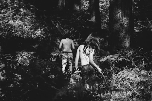 A black and white photograph of two children walking away into a dense forest. Both are bare-backed with patterns painted on their backs, surrounded by tall trees and ferns. The light and shadow interplay creates a mysterious and adventurous atmosphere.