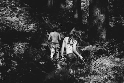 A black and white photograph of two children walking away into a dense forest. Both are bare-backed with patterns painted on their backs, surrounded by tall trees and ferns. The light and shadow interplay creates a mysterious and adventurous atmosphere.