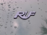 A glossy emblem displays the letters 'RUF' against a green metallic surface dotted with water droplets, indicating a recent rain or a moist environment.
