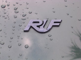 A glossy emblem displays the letters 'RUF' against a green metallic surface dotted with water droplets, indicating a recent rain or a moist environment.