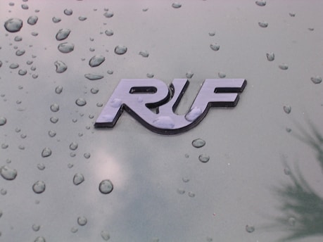 A glossy emblem displays the letters 'RUF' against a green metallic surface dotted with water droplets, indicating a recent rain or a moist environment.