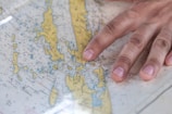 A close-up of a hand placed on a nautical map featuring yellow islands with blue water regions surrounding them. The hand is positioned over a central island, suggesting an act of navigation or orientation. The map includes various labels and symbols denoting geological and navigational information.