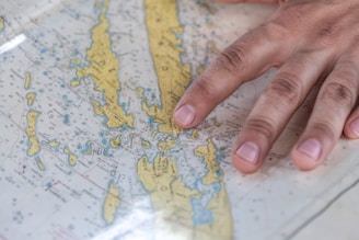 A close-up of a hand placed on a nautical map featuring yellow islands with blue water regions surrounding them. The hand is positioned over a central island, suggesting an act of navigation or orientation. The map includes various labels and symbols denoting geological and navigational information.