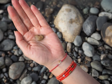A hand holds a small heart-shaped stone over a surface covered with variously colored pebbles. The hand is adorned with red beaded bracelets, adding a pop of color.