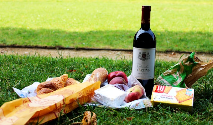 A picnic setup on grass with a bottle of red wine, croissants, baguette, cheese, peaches, and a packet of biscuits.