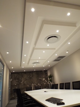A modern conference room features a long white table surrounded by black chairs. The ceiling is adorned with recessed lighting and circular air conditioning vents. A patterned accent wall and a potted plant enhance the room’s decor.