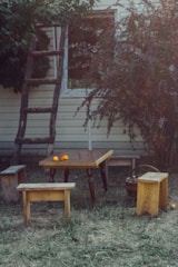 A rustic outdoor setting featuring a small wooden table with two matching benches and an additional stool. The table has a few small orange flowers placed on it. In the background, there is an old wooden ladder leaning against a house with a window partially obscured by foliage from overgrown bushes.