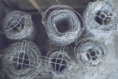 Coils of barbed wire are stacked on top of each other on a concrete surface. The rolls are tightly wound and secured with additional wire. The setting appears to be an industrial or storage area, possibly a warehouse or workshop.