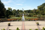 A well-manicured garden featuring a symmetrical layout with a central water feature. Bright flowers in shades of orange and yellow line the paths, surrounded by lush greenery and neatly trimmed bushes. Tall trees frame the garden, and a spacious paved area with potted plants is in the foreground. The sky is clear and blue, enhancing the serene atmosphere.