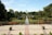 A well-manicured garden featuring a symmetrical layout with a central water feature. Bright flowers in shades of orange and yellow line the paths, surrounded by lush greenery and neatly trimmed bushes. Tall trees frame the garden, and a spacious paved area with potted plants is in the foreground. The sky is clear and blue, enhancing the serene atmosphere.