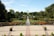 A well-manicured garden featuring a symmetrical layout with a central water feature. Bright flowers in shades of orange and yellow line the paths, surrounded by lush greenery and neatly trimmed bushes. Tall trees frame the garden, and a spacious paved area with potted plants is in the foreground. The sky is clear and blue, enhancing the serene atmosphere.