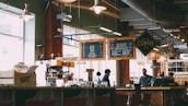 A cozy coffee shop with two individuals behind the counter preparing drinks. Overhead menu boards display various coffee options. The interior has a warm and inviting ambiance with soft lighting from hanging lamps. Wooden furniture and shelves filled with coffee-related items add to the rustic charm.
