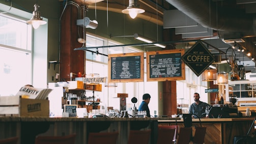 A cozy coffee shop with two individuals behind the counter preparing drinks. Overhead menu boards display various coffee options. The interior has a warm and inviting ambiance with soft lighting from hanging lamps. Wooden furniture and shelves filled with coffee-related items add to the rustic charm.