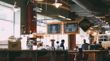 A cozy coffee shop with two individuals behind the counter preparing drinks. Overhead menu boards display various coffee options. The interior has a warm and inviting ambiance with soft lighting from hanging lamps. Wooden furniture and shelves filled with coffee-related items add to the rustic charm.