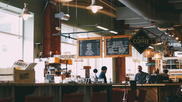 A cozy coffee shop with two individuals behind the counter preparing drinks. Overhead menu boards display various coffee options. The interior has a warm and inviting ambiance with soft lighting from hanging lamps. Wooden furniture and shelves filled with coffee-related items add to the rustic charm.