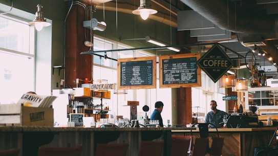 A cozy coffee shop with two individuals behind the counter preparing drinks. Overhead menu boards display various coffee options. The interior has a warm and inviting ambiance with soft lighting from hanging lamps. Wooden furniture and shelves filled with coffee-related items add to the rustic charm.