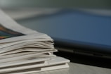 A stack of newspapers lies on a light-colored surface, with some pages slightly curling at the edges. Nearby, part of a laptop or similar electronic device is visible, suggesting a juxtaposition of print media and digital technology.
