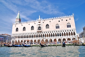 A grand historic palace with intricate gothic architecture stands prominently behind a canal filled with gondolas. The building features ornate windows, a lattice pattern on its facade, and a distinct tower with a spire. The sky is clear and blue, and visitors gather around the waterfront, adding a lively atmosphere.