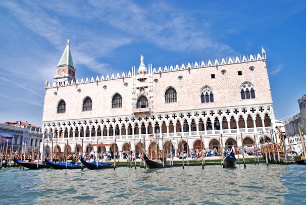 A grand historic palace with intricate gothic architecture stands prominently behind a canal filled with gondolas. The building features ornate windows, a lattice pattern on its facade, and a distinct tower with a spire. The sky is clear and blue, and visitors gather around the waterfront, adding a lively atmosphere.