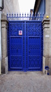 A vibrant blue wrought iron gate with ornate patterns, framed by stone pillars. There is a metal sign on the gate and a small object resembling a street number plate on one pillar. The pavement is cobblestone and the gate leads to what appears to be a narrow alleyway or side street. The top of the gate features pointed finials.