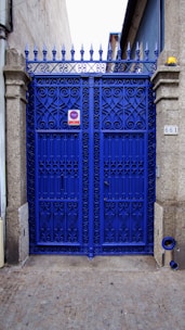 A vibrant blue wrought iron gate with ornate patterns, framed by stone pillars. There is a metal sign on the gate and a small object resembling a street number plate on one pillar. The pavement is cobblestone and the gate leads to what appears to be a narrow alleyway or side street. The top of the gate features pointed finials.