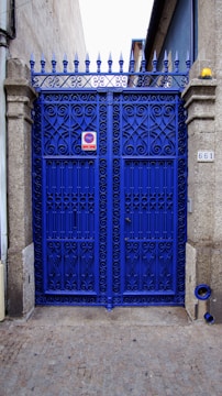 A vibrant blue wrought iron gate with ornate patterns, framed by stone pillars. There is a metal sign on the gate and a small object resembling a street number plate on one pillar. The pavement is cobblestone and the gate leads to what appears to be a narrow alleyway or side street. The top of the gate features pointed finials.