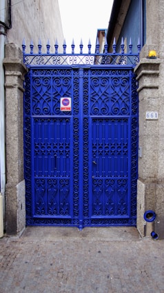 A vibrant blue wrought iron gate with ornate patterns, framed by stone pillars. There is a metal sign on the gate and a small object resembling a street number plate on one pillar. The pavement is cobblestone and the gate leads to what appears to be a narrow alleyway or side street. The top of the gate features pointed finials.