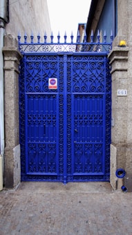 A vibrant blue wrought iron gate with ornate patterns, framed by stone pillars. There is a metal sign on the gate and a small object resembling a street number plate on one pillar. The pavement is cobblestone and the gate leads to what appears to be a narrow alleyway or side street. The top of the gate features pointed finials.