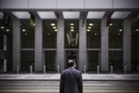 A man in a suit stands on the street facing a large, modern building with a glass and concrete facade. The atmosphere is urban and somewhat subdued, with muted lighting and minimal activity.
