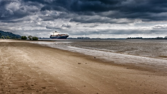 A large cargo ship sails on the water near a deserted sandy beach. The sky is overcast with dark, heavy clouds, creating a dramatic and moody atmosphere. In the distance, industrial buildings are visible along the shoreline, surrounded by trees and hills.