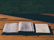 An open book and a notebook with handwritten notes sit on a wooden table under natural light. A pencil is placed in the book, suggesting it's being used for study or preparation.