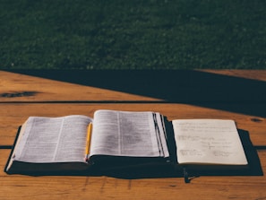 An open book and a notebook with handwritten notes sit on a wooden table under natural light. A pencil is placed in the book, suggesting it's being used for study or preparation.